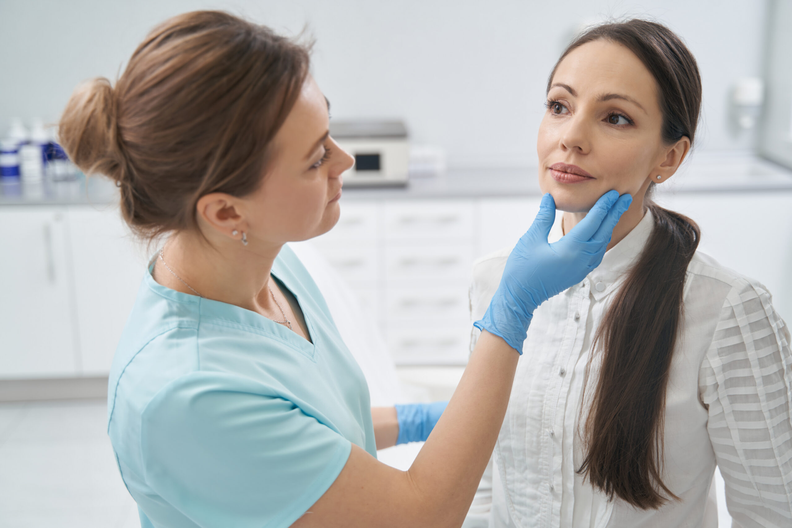 Medical clinician in blue gloves gently supports a patient's chin during a checkup in a bright clinic room.
